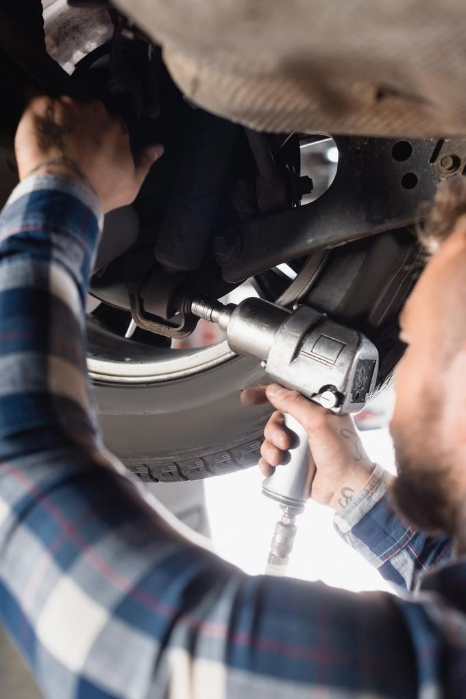Un mecánico utiliza una llave eléctrica para reparar la parte inferior de un coche, mientras otra persona sostiene las piezas cerca.