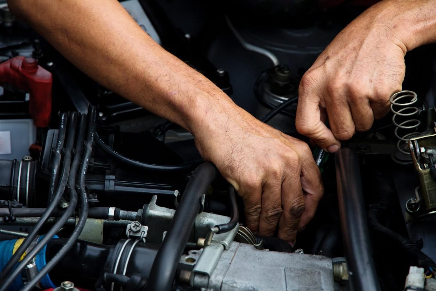 Manos de mecánico trabajando dentro del compartimento del motor de un automóvil con herramientas y cableado expuesto.