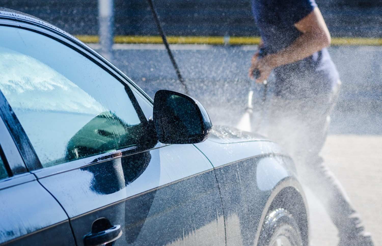 Una persona lava un coche azul oscuro con una hidrolavadora; el agua salpica la calle.