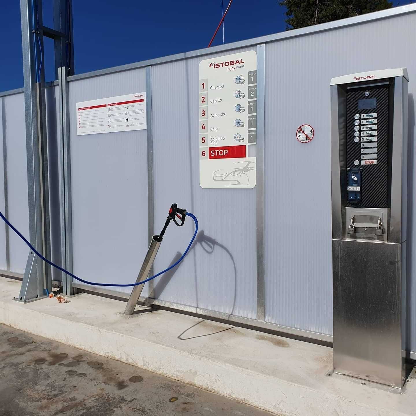 Estación de servicios exteriores con manguera y panel de control junto a una valla metálica gris, al lado de una máquina expendedora plateada.
