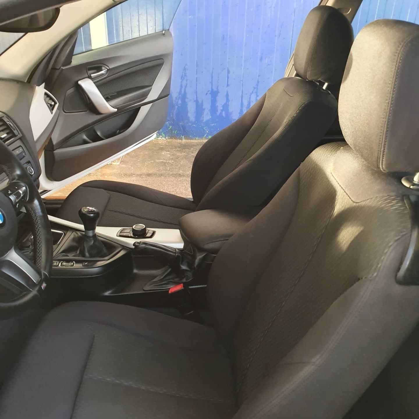 Interior del coche con dos asientos delanteros negros, palanca de cambios manual y volante visibles.