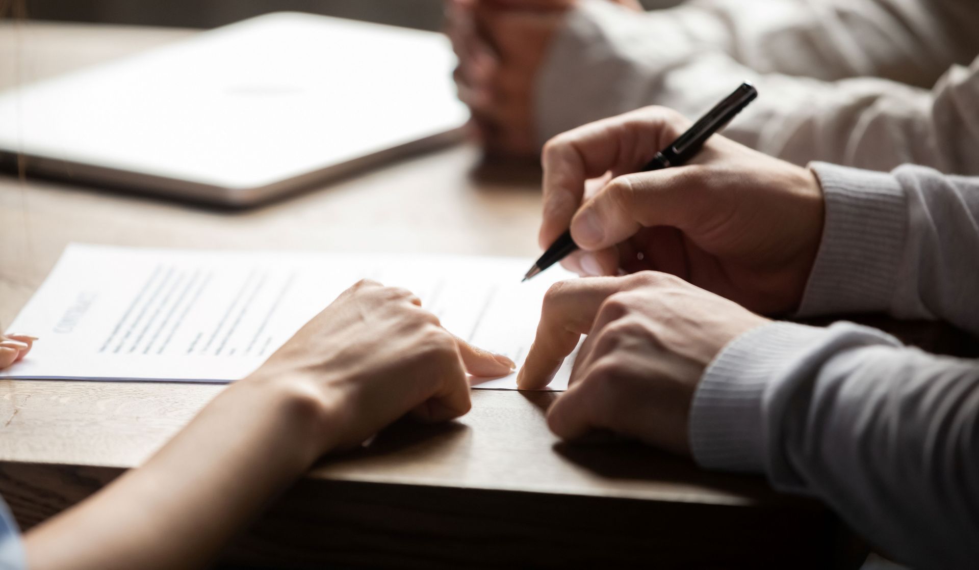 Des mains écrivent sur du papier à une table en bois, avec un ordinateur portable en arrière-plan