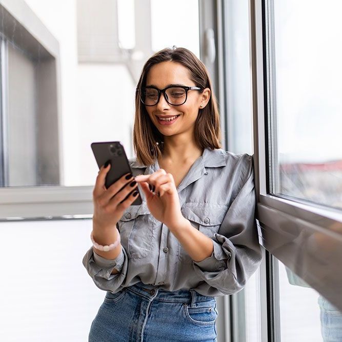 Eine Frau mit Brille lehnt an einem Fenster und schaut auf ihr Mobiltelefon.