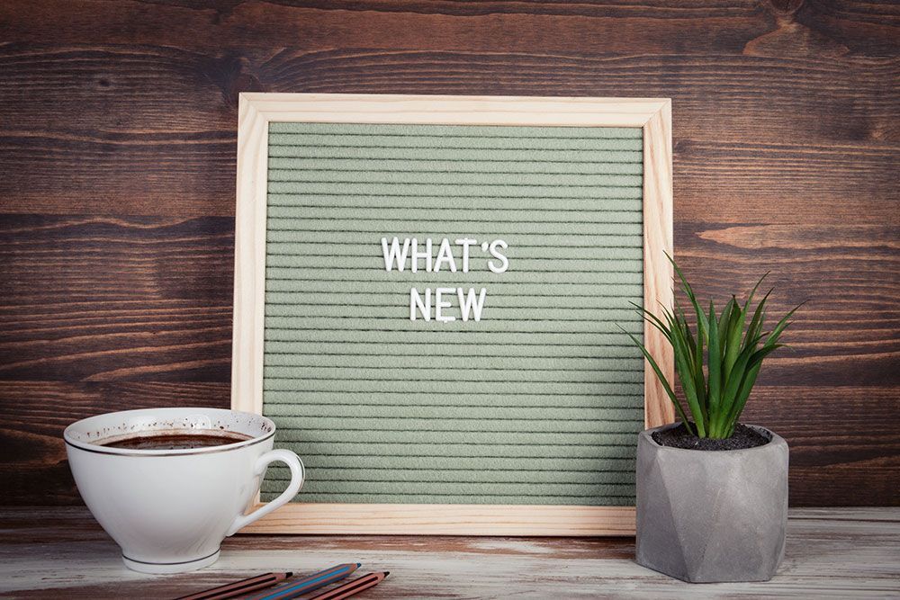 Eine Buchstabentafel mit der Aufschrift „Was gibt’s Neues“ neben einer Tasse Kaffee und einer Topfpflanze.