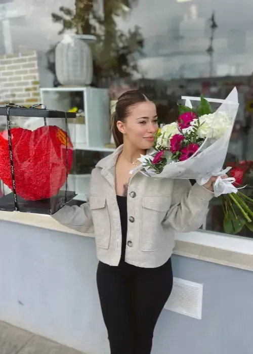 Mujer sosteniendo un ramo, de pie frente a una tienda con flores y un arreglo de corazones.