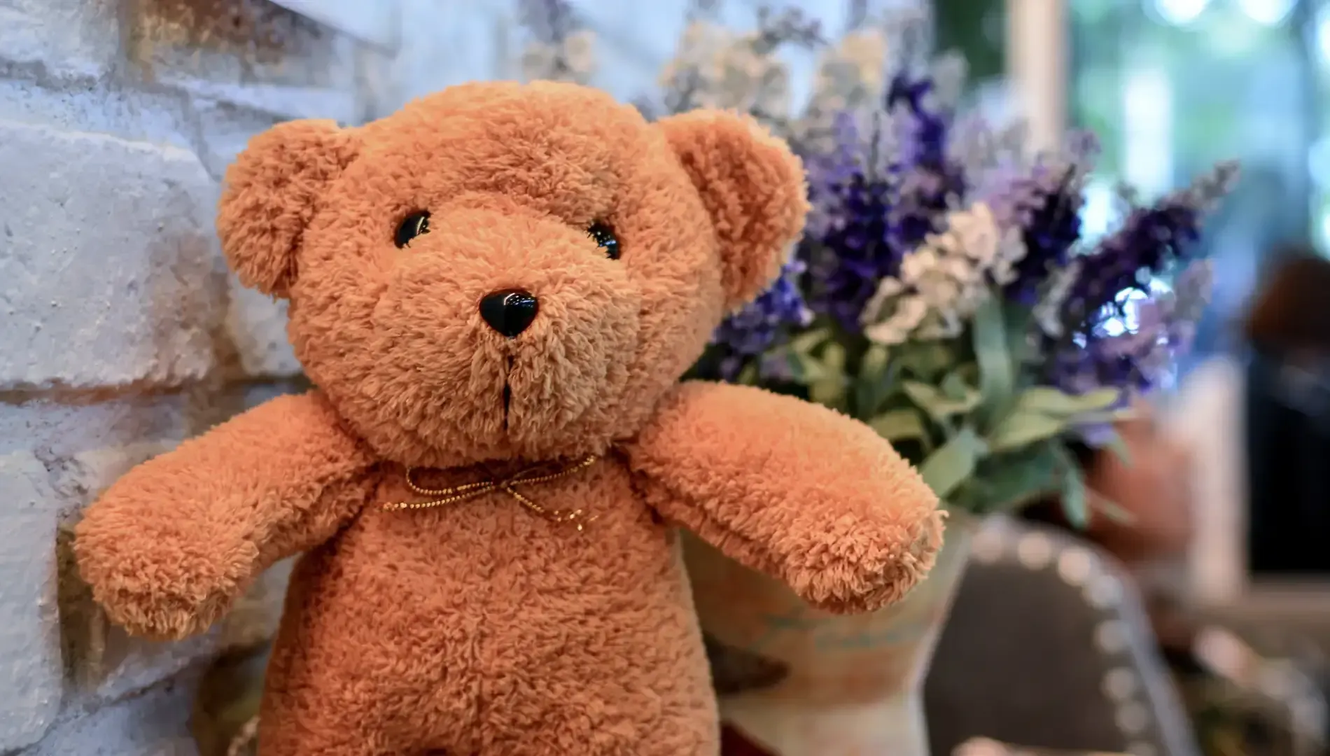 Osito de peluche marrón con ojos negros y collar dorado, delante de flores moradas en un jarrón.