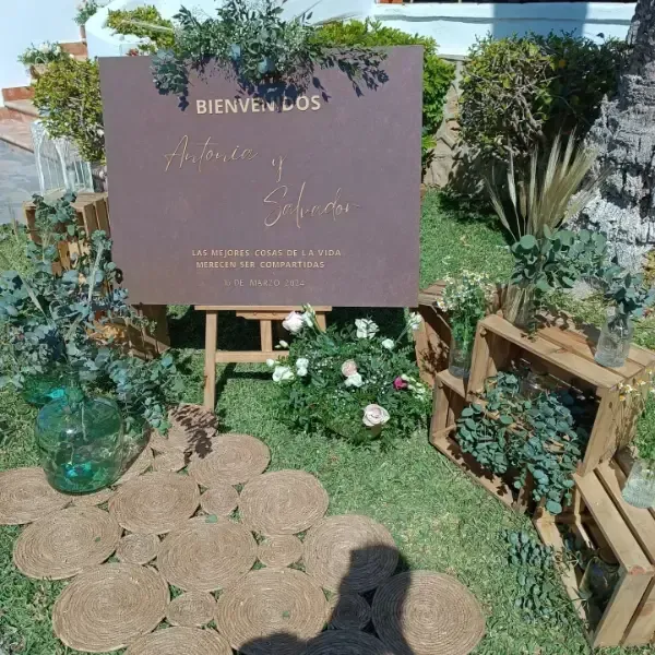 Cartel de bienvenida de boda en un jardín, cartel marrón con nombres y escritura, decorado con flores y vegetación.