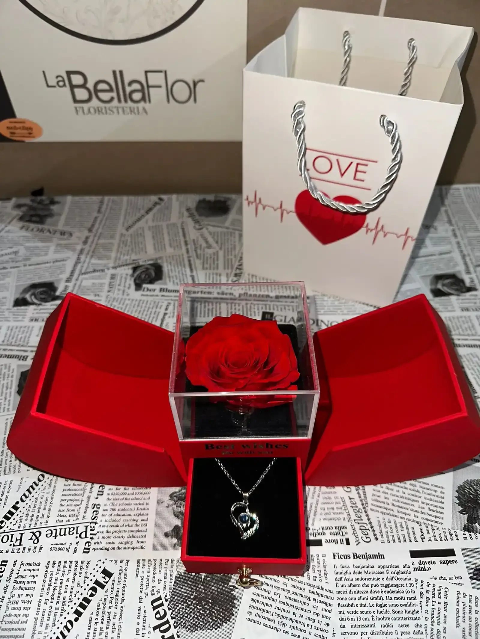 Rosa roja en un joyero con collar y bolsa de regalo, presentada sobre un fondo de periódico.