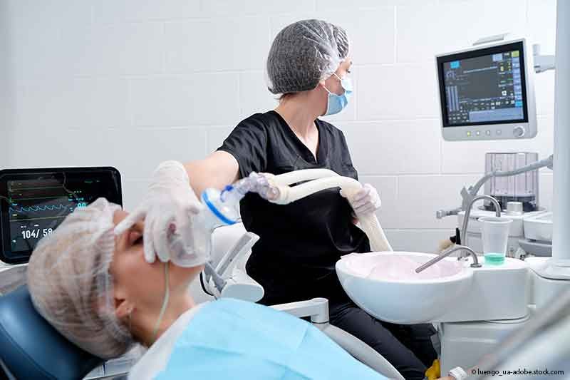 Zahnarzt verabreicht einem Patienten im Zahnarztstuhl ein Beruhigungsmittel; Monitore und Geräte sind sichtbar.