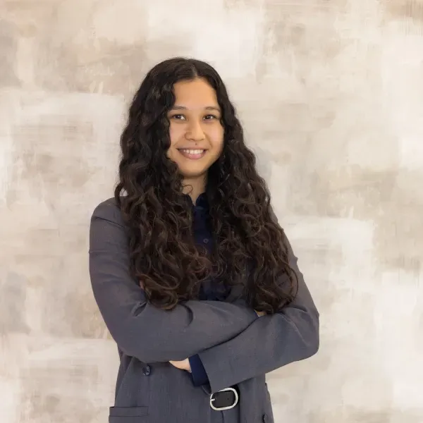 Una mujer con cabello largo y rizado está de pie con los brazos cruzados y sonriendo.