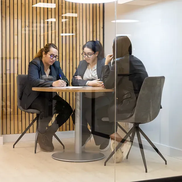 Tres personas están sentadas en una mesa detrás de una pared de cristal.