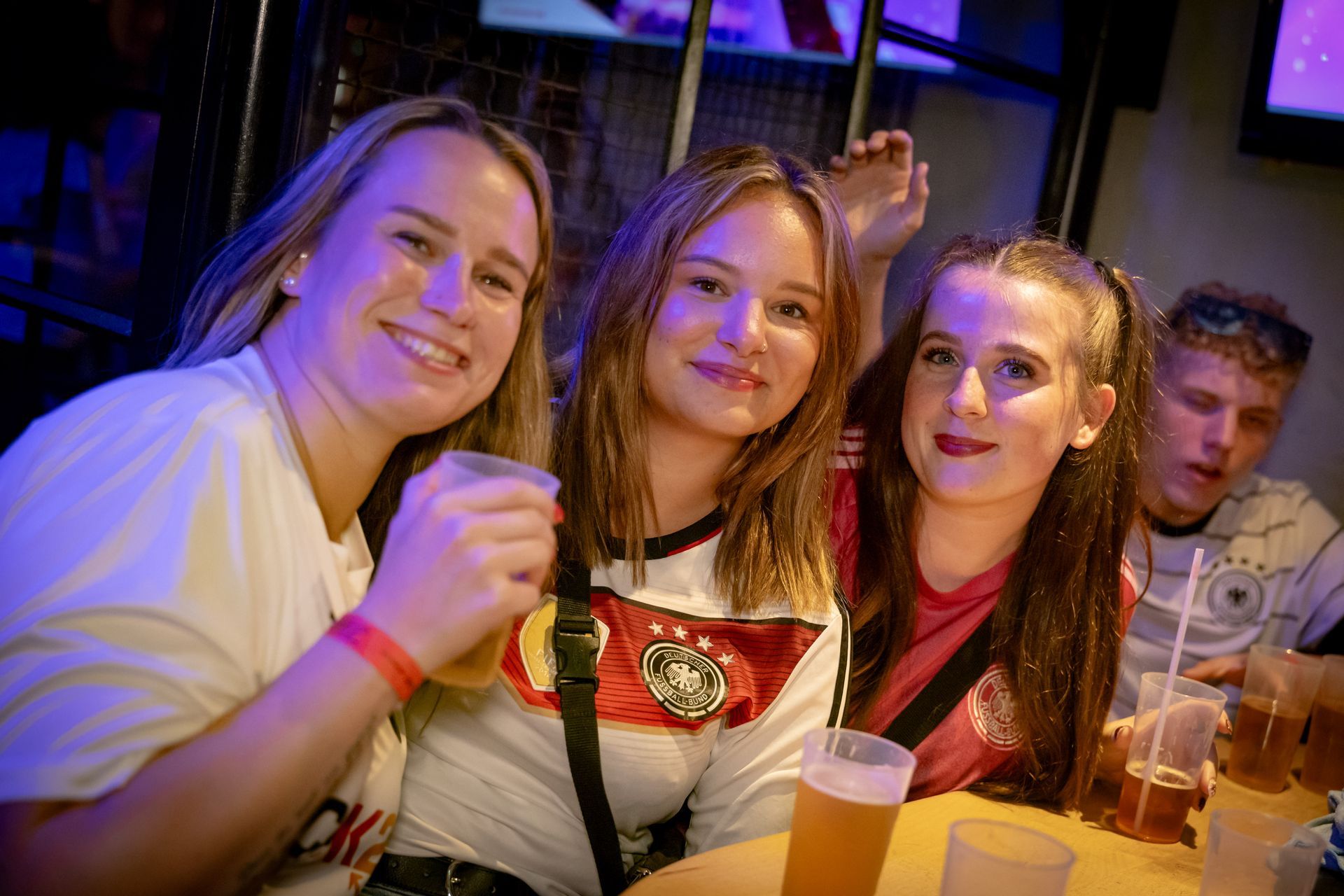 Drei lächelnde Personen mit Getränken in der Hand auf einer schwach beleuchteten Party oder in einer Bar.