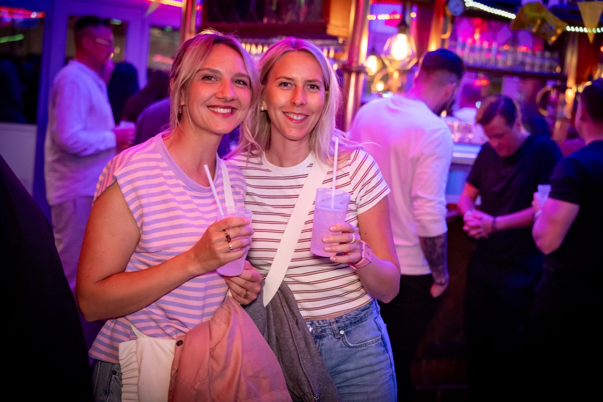 Zwei Frauen in gestreiften Oberteilen halten Getränke und lächeln in einer farbenfrohen Barszene