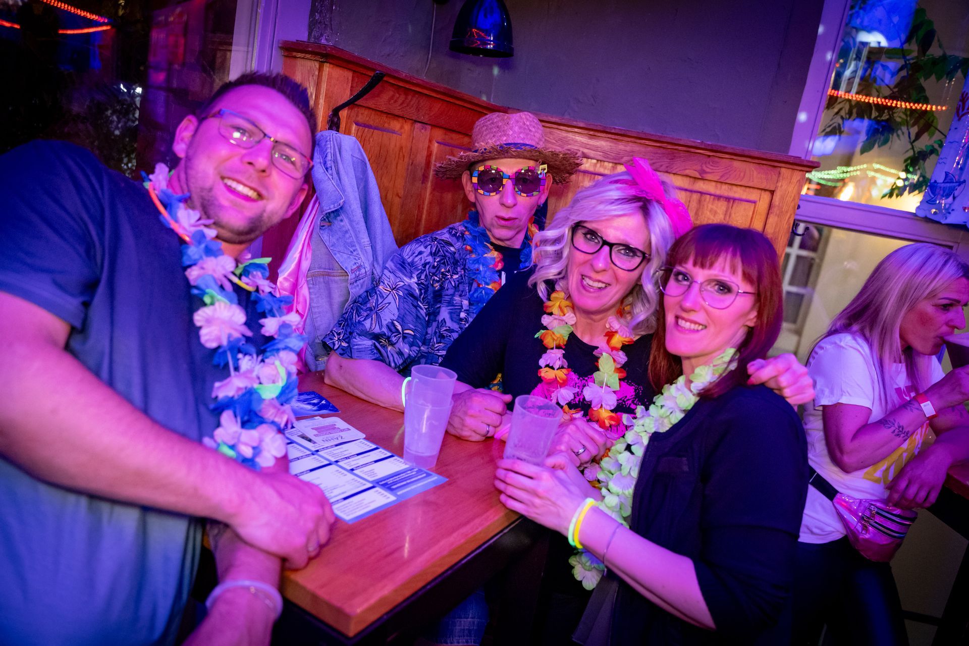 Vier Freundinnen lächeln an einem bunten Nachtclubtisch, sie tragen Blumenkränze und einen Hut.