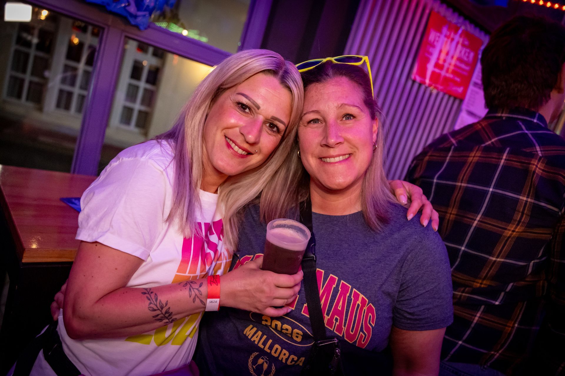 Zwei lächelnde und posierende Personen an einer Bar, die Getränke unter rosa und violettem Licht in den Händen.