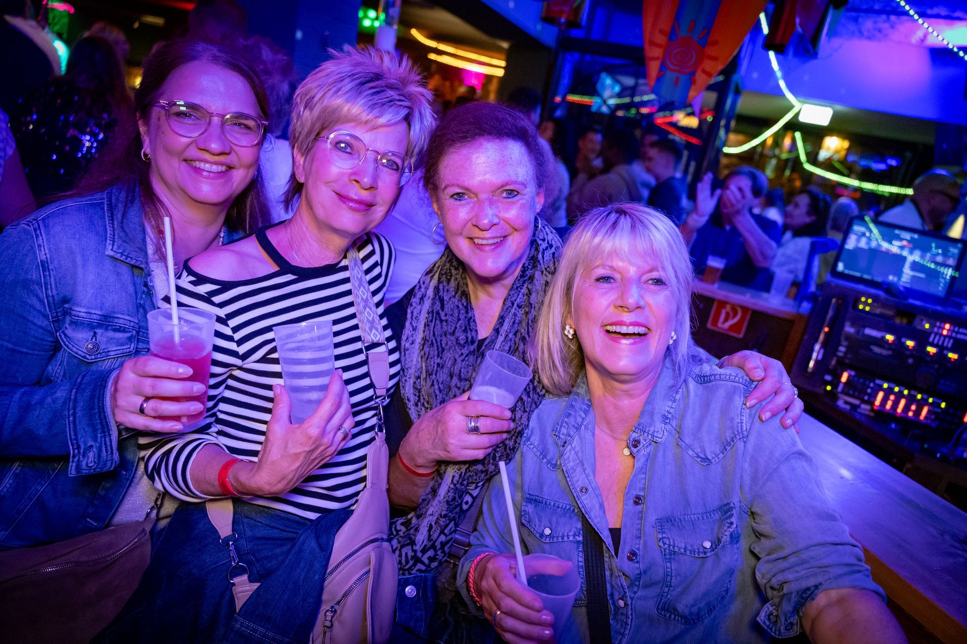 Vier lächelnde Frauen mit Getränken in der Hand posieren gemeinsam an einem Tisch in einem neonbeleuchteten Nachtclub.