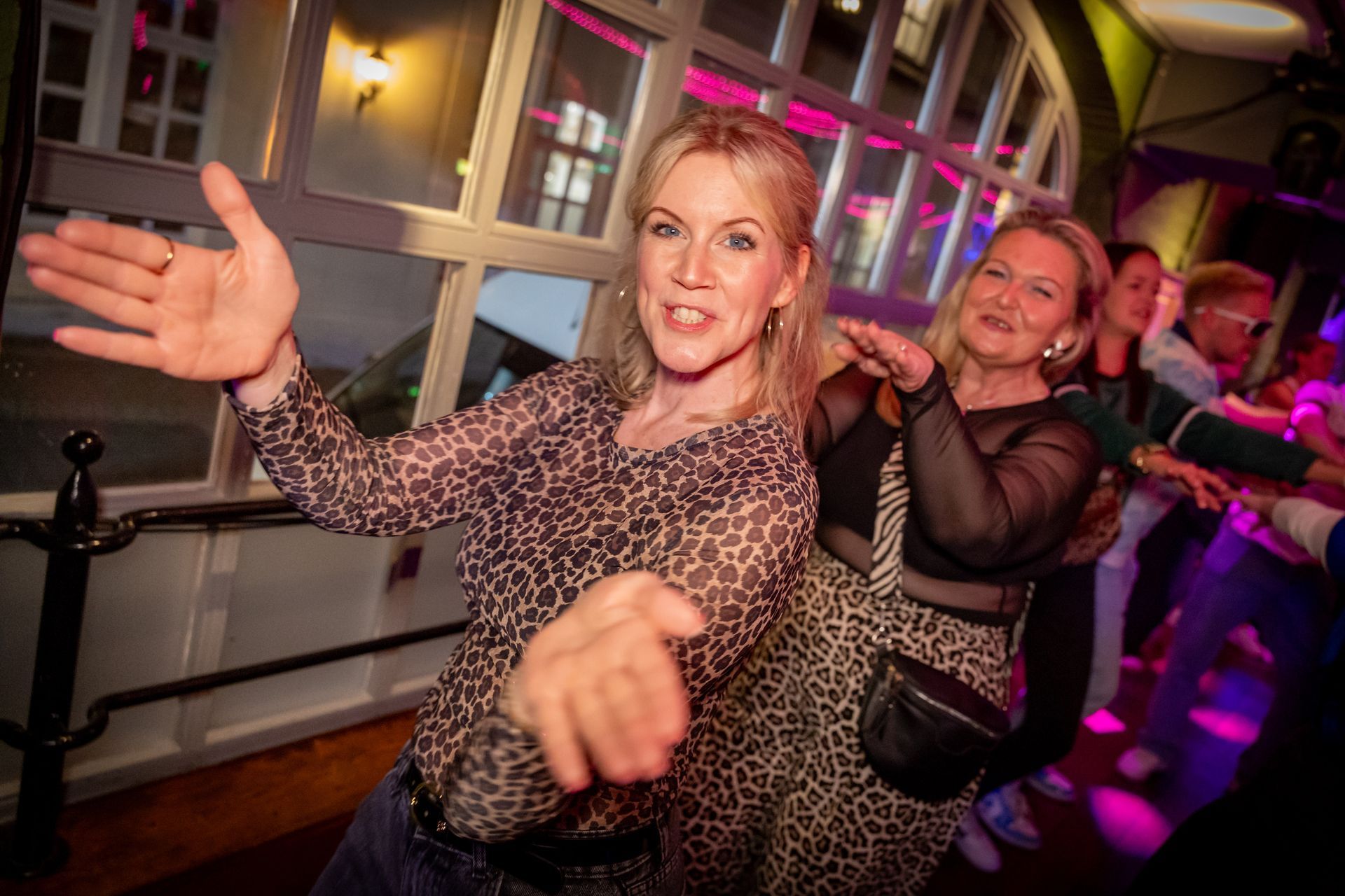 Zwei Frauen tanzen auf einer ausgelassenen Party, eine streckt lächelnd die Hand in Richtung Kamera aus; im Hintergrund sind violette Lichter zu sehen.