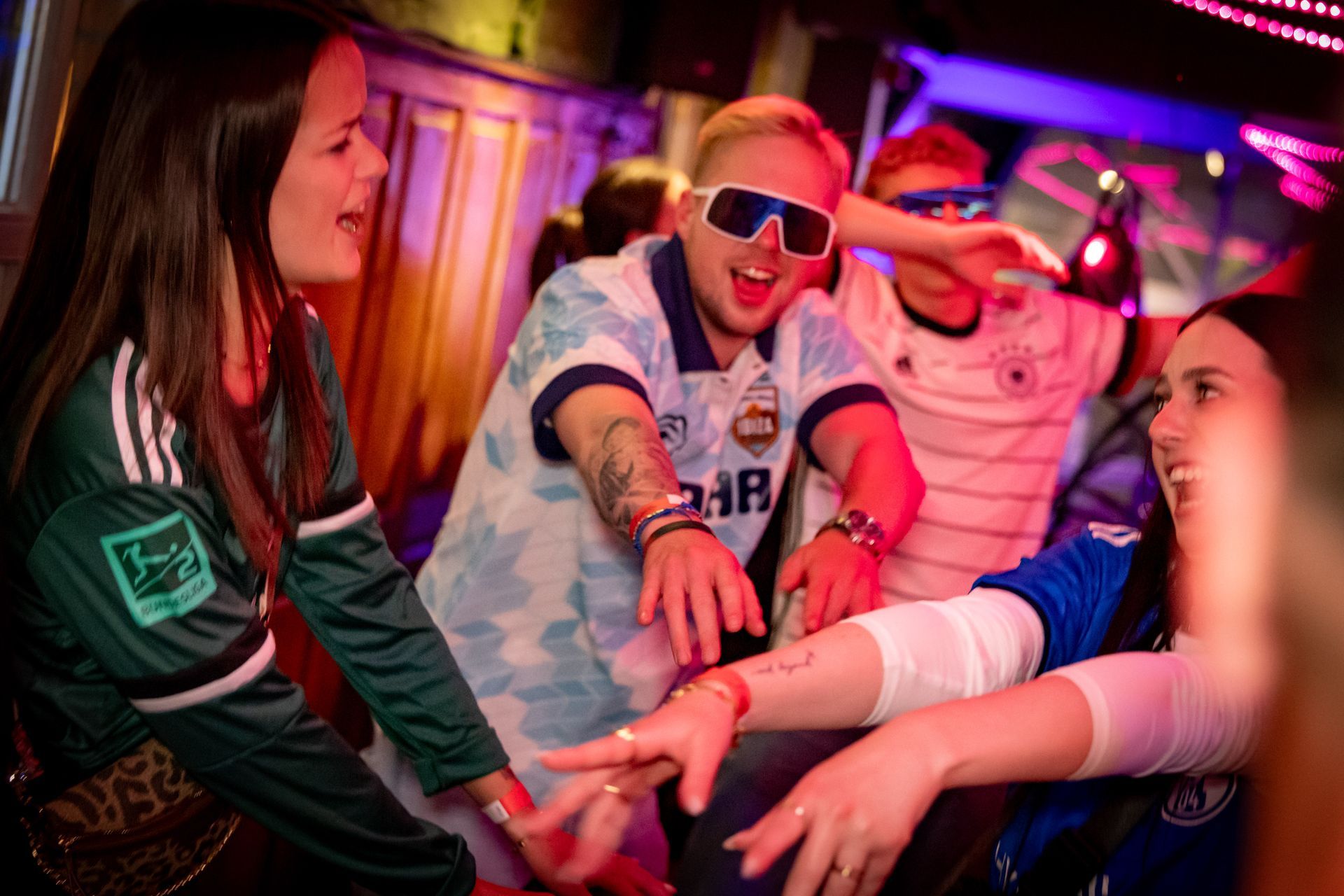 Freunde greifen sich unter Neonlichtern auf einer ausgelassenen Party die Hände, lächeln und jubeln.