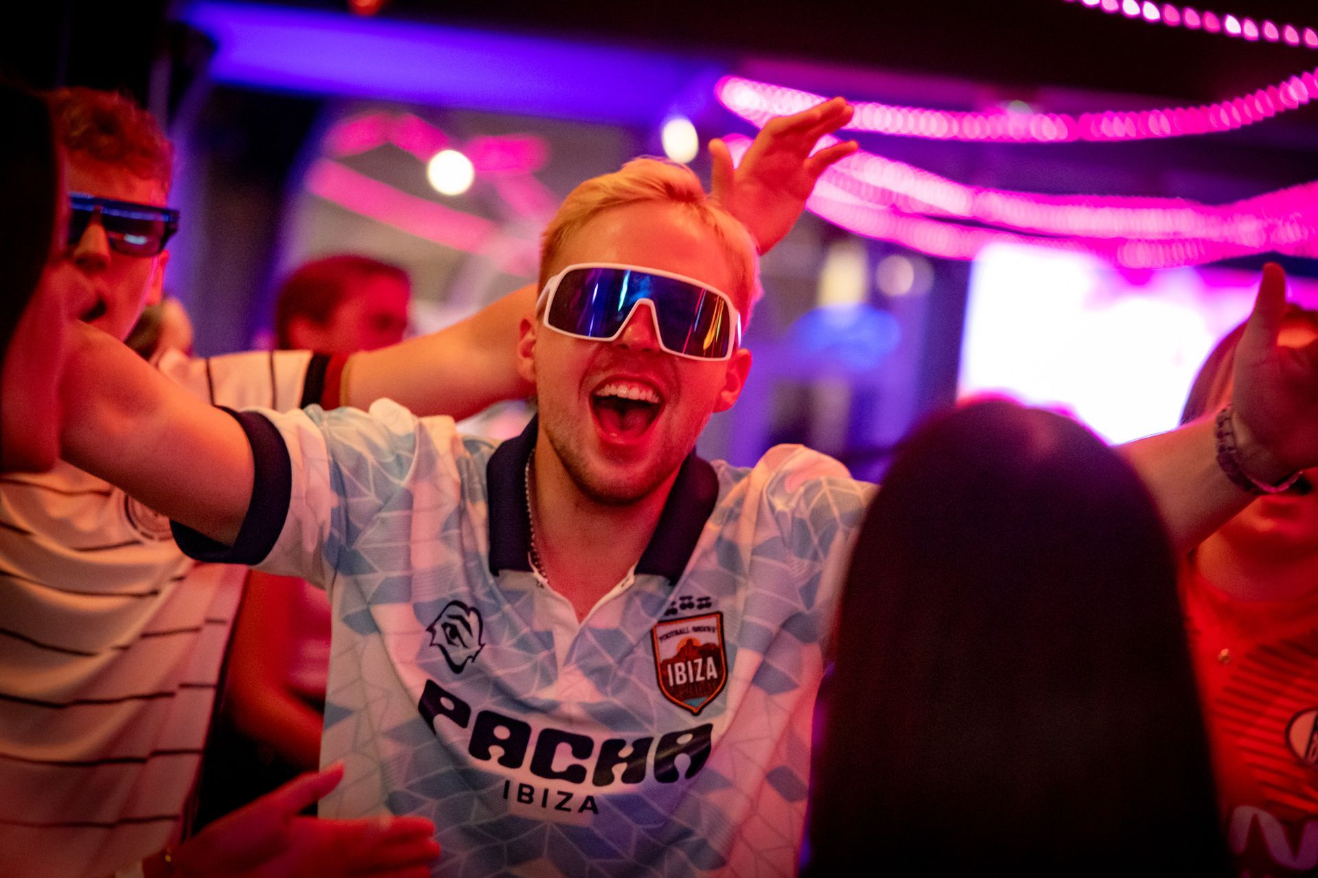 In einem neonbeleuchteten Nachtclub jubelt eine Menge; ein Mann mit verspiegelter Sonnenbrille lächelt und hebt die Arme.
