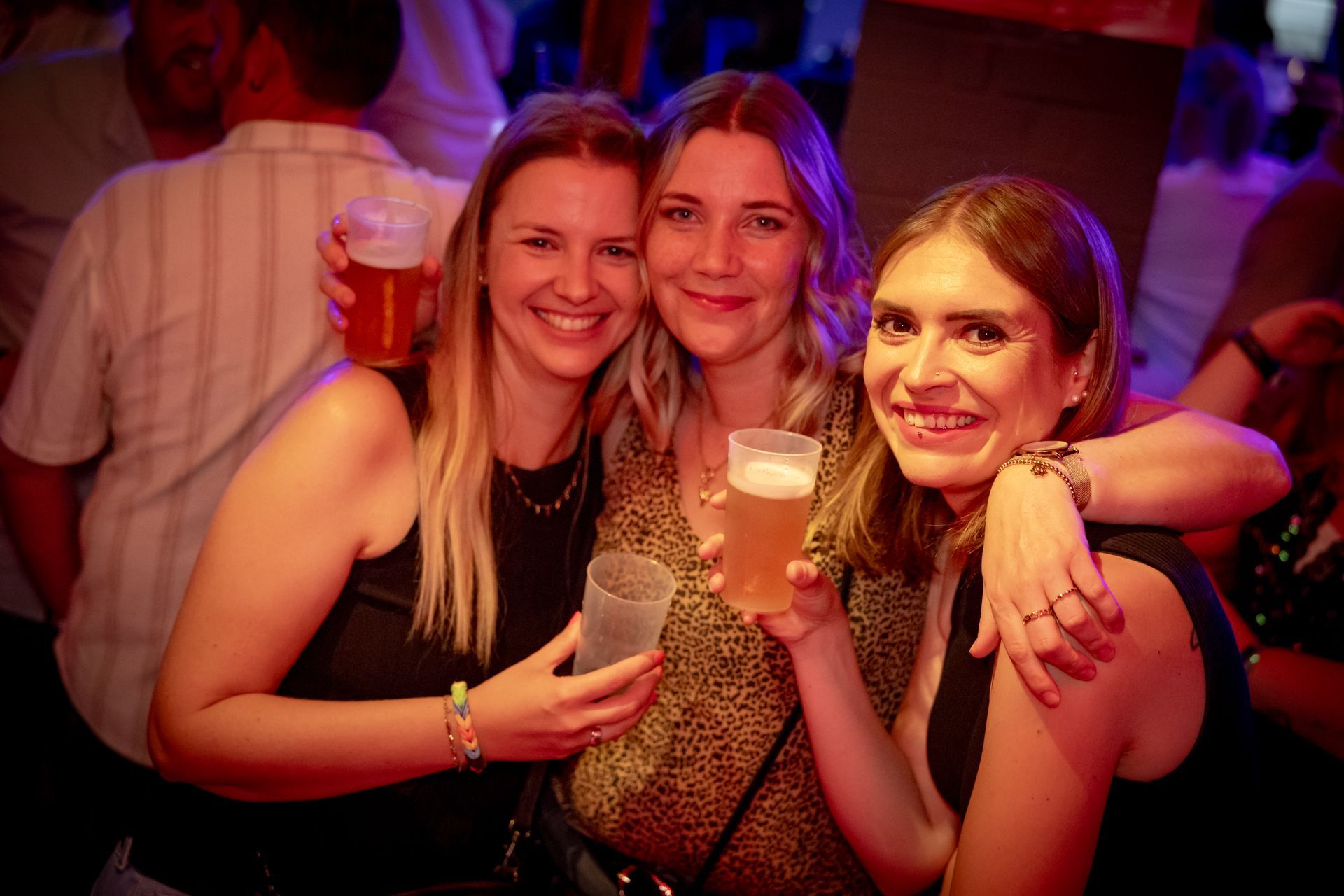 Drei Freunde lächeln und halten Getränke in der Hand auf einer schwach beleuchteten Party.
