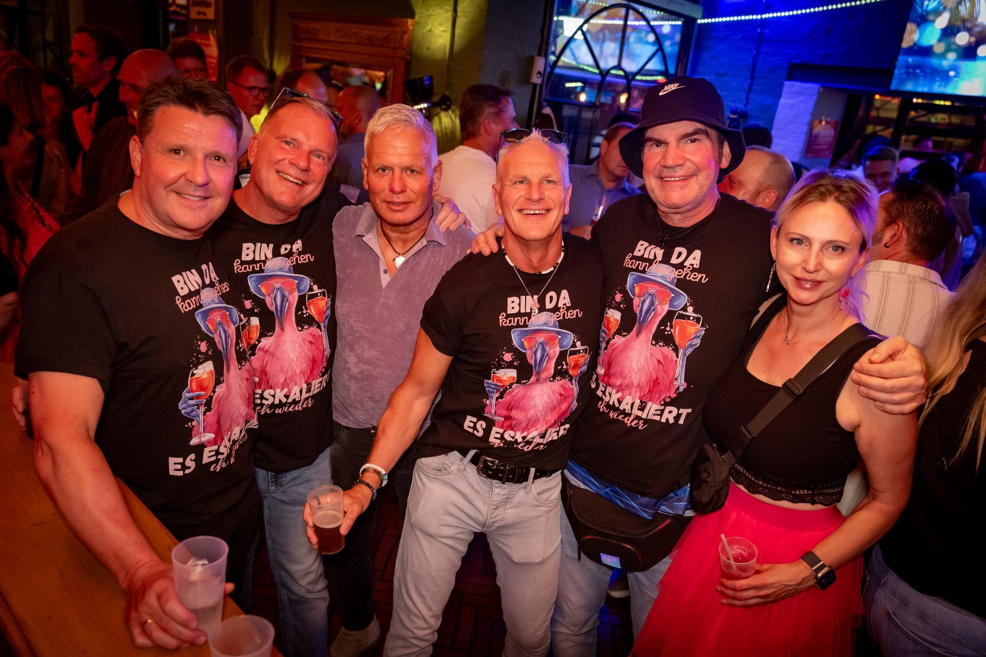 Sechs lächelnde Menschen auf einer ausgelassenen Party mit bunter Beleuchtung und festlichen Hemden.