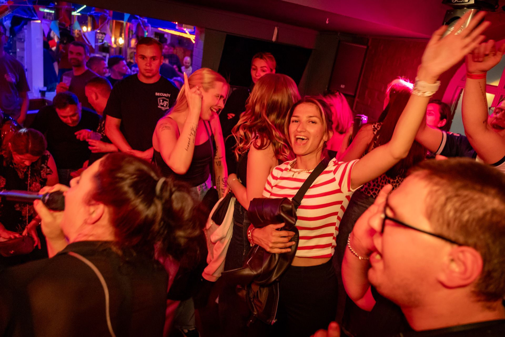 Eine Menschenmenge tanzt in einem neonbeleuchteten Nachtclub; die Leute lächeln und heben die Arme.