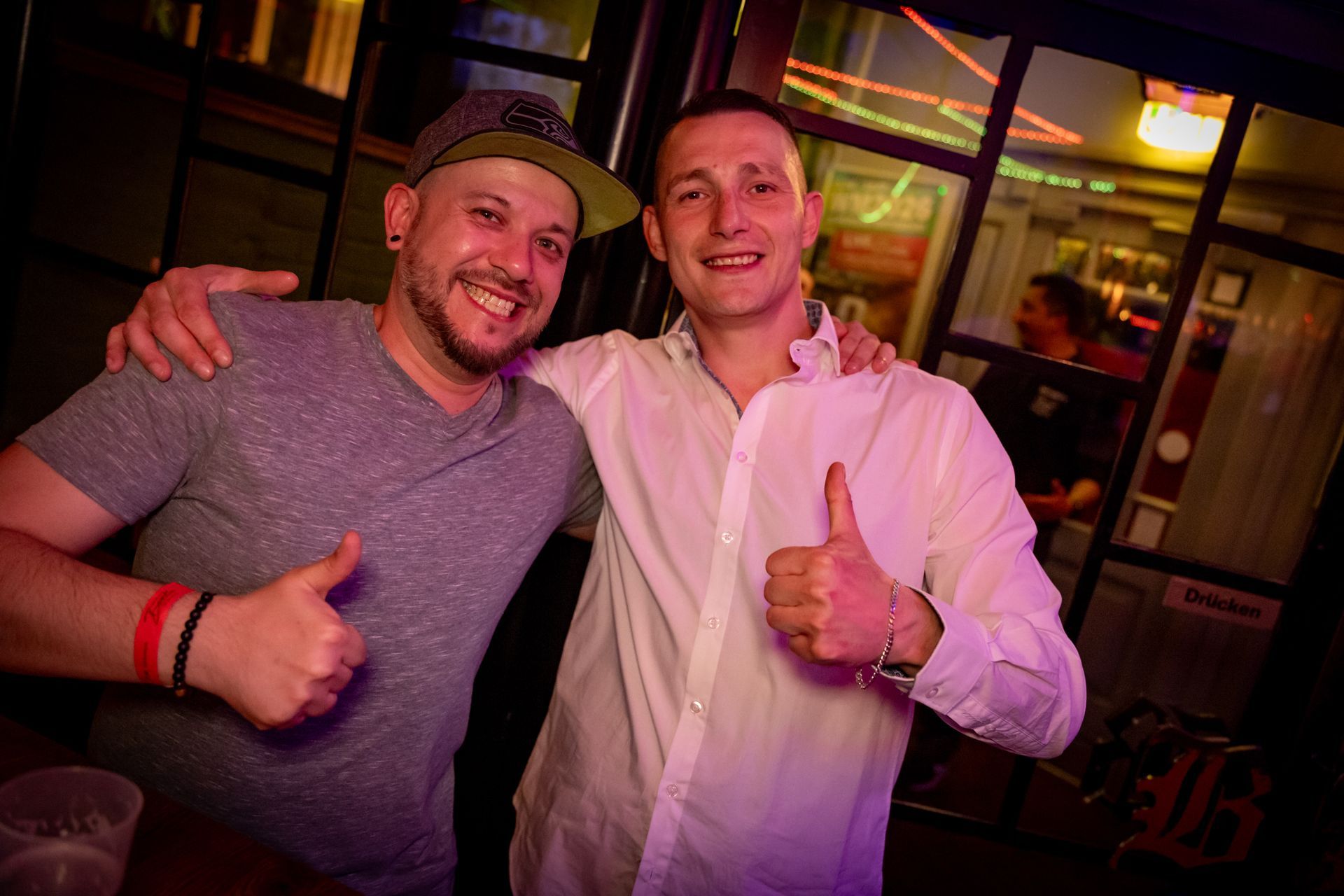 Zwei lächelnde Männer in einer Bar, die eng beieinander stehen und sich gegenseitig den Daumen hoch zeigen.