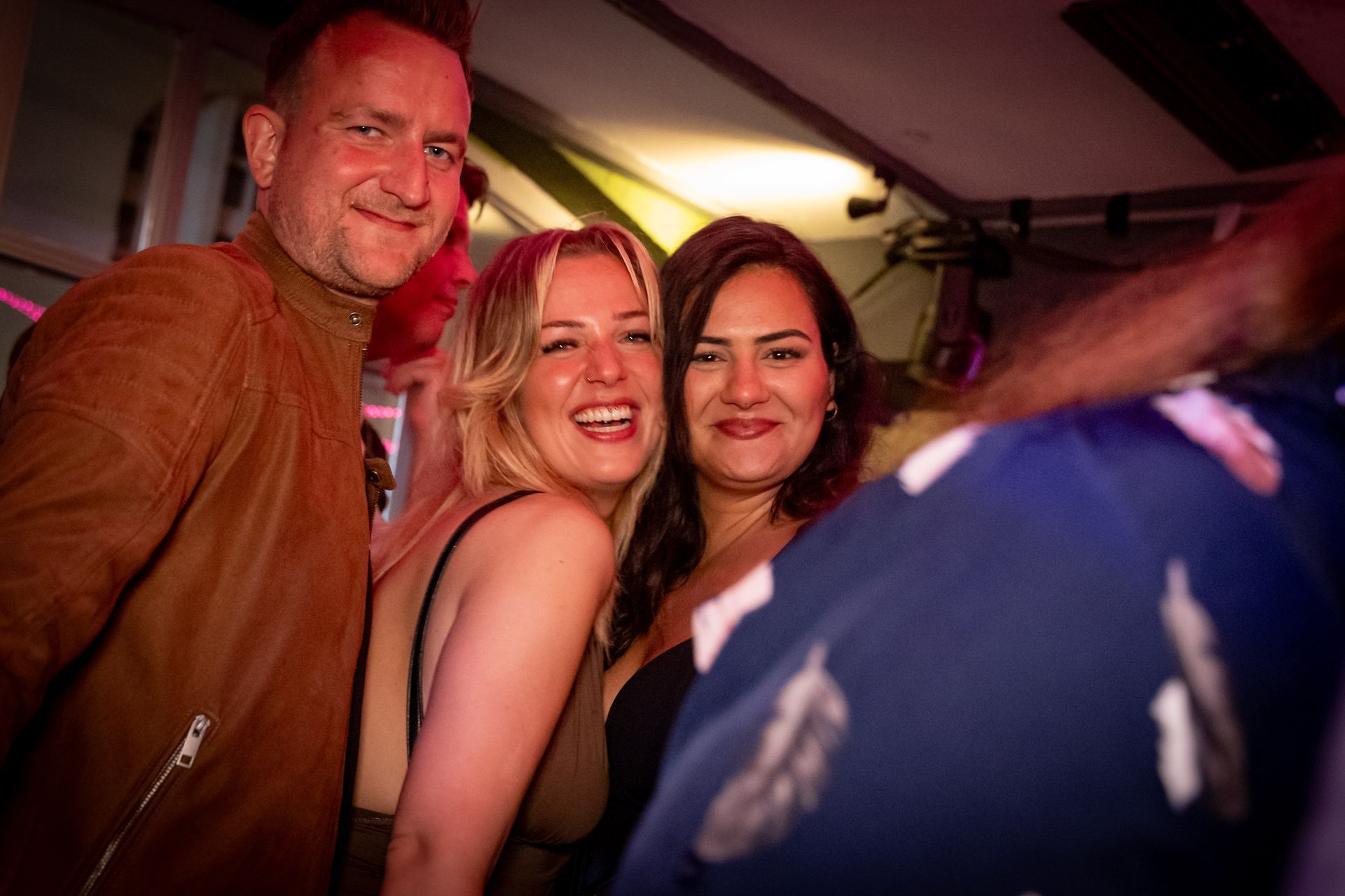 Drei lächelnde Personen auf einer Party, warmes rosa Licht, im Vordergrund eine verschwommene Person.