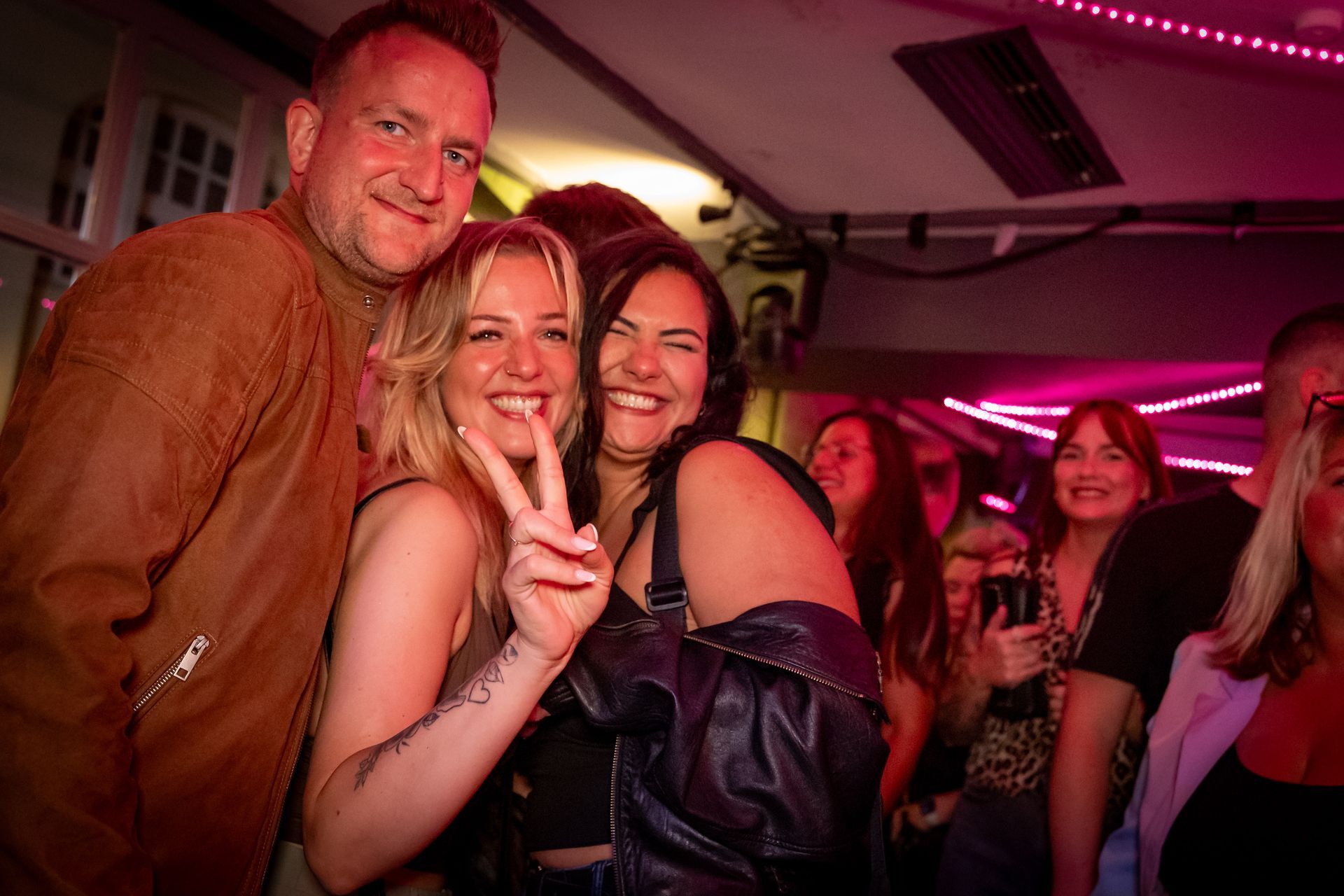 Drei lächelnde Personen posieren in einem rosa beleuchteten Partybus, wobei eine von ihnen ein Friedenszeichen zeigt.