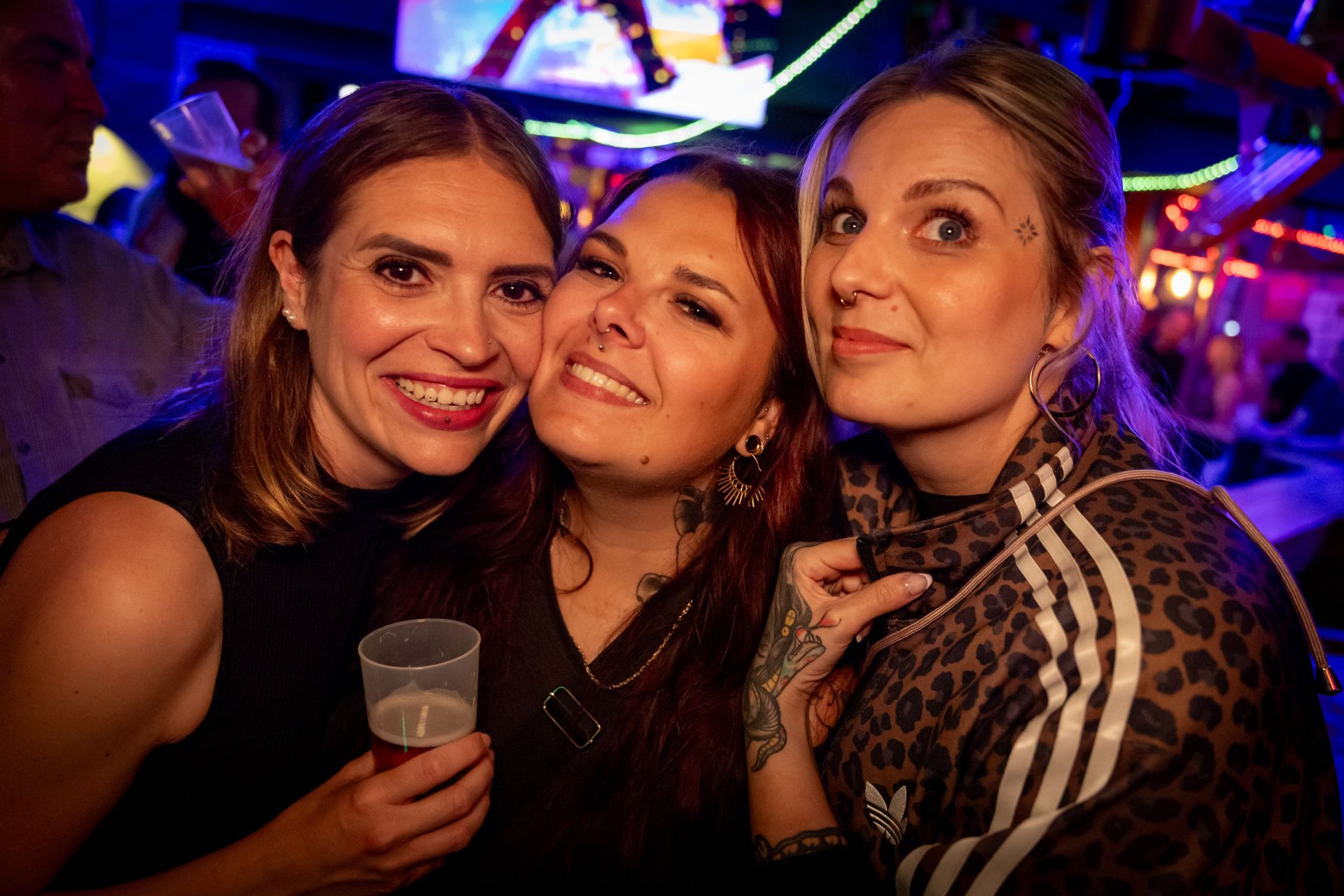 Drei Freunde lächeln in einem Nachtclub und halten unter bunten Lichtern ein Getränk in der Hand.