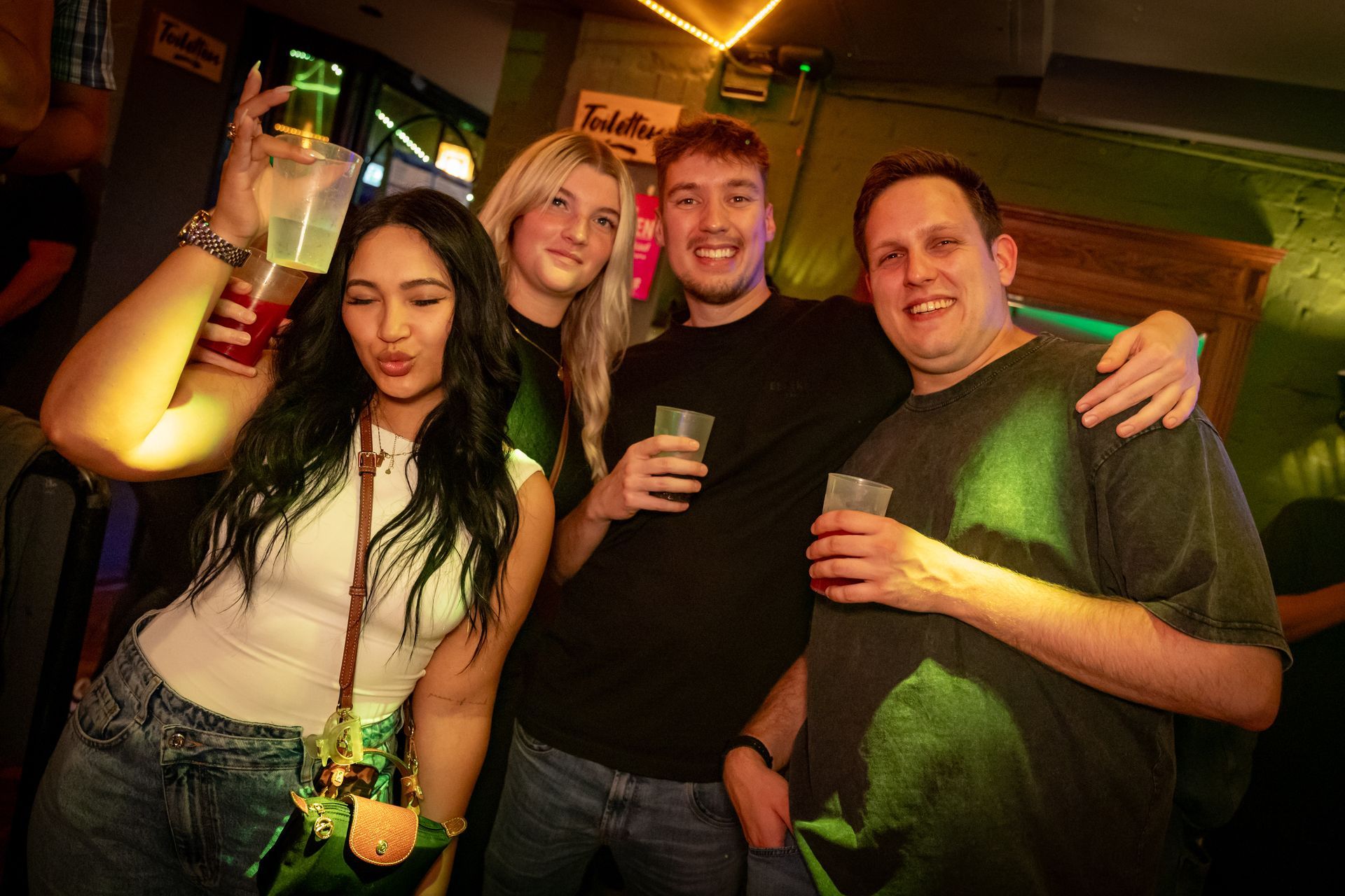 Vier Freunde lächeln und posieren mit Getränken in einer schwach beleuchteten Bar.