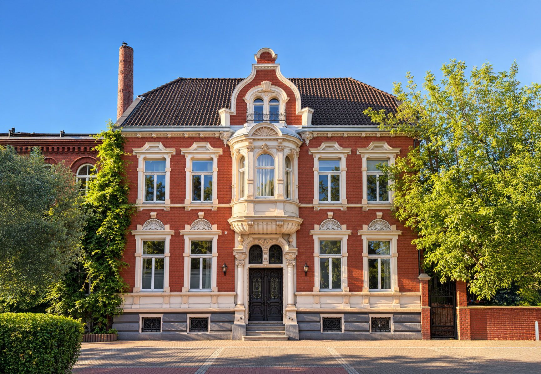 Prächtiges historisches Gebäude aus rotem Backstein mit weißen Verzierungen und einem zentralen Eingang, eingerahmt von Bäumen