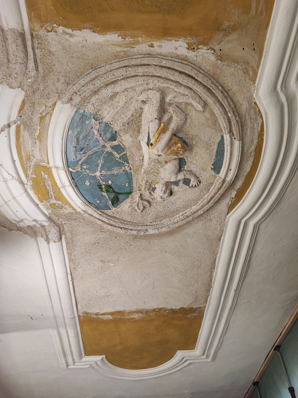 Eine verwitterte, ovale Reliefskulptur eines Vogels, die in eine gold-weiße Gipsleiste an einer Decke eingelassen ist.