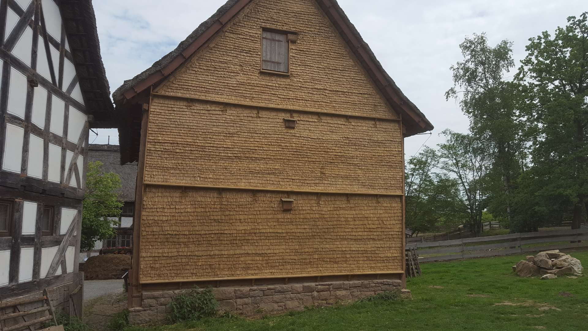 Seitenansicht eines Hauses mit braunen, strohgedeckten Wänden und Steinfundament, neben einem schwarz-weißen Fachwerkgebäude.