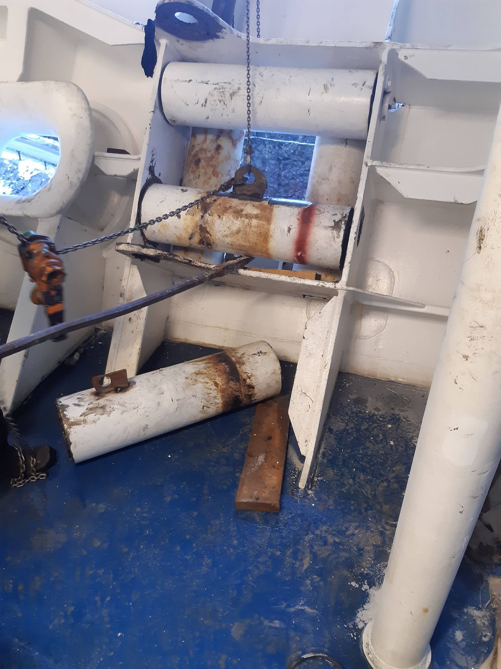 Ruosteinen laivankansi, jossa valkoiset putket, sininen lattia ja valtameri näkyy pyöreästä ikkunasta.