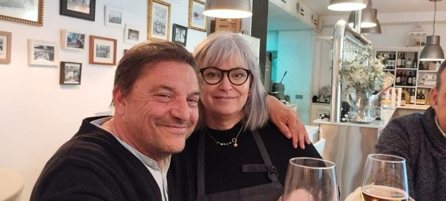 Dos personas sonriendo en una mesa de restaurante con copas de vino y fotos enmarcadas en la pared.