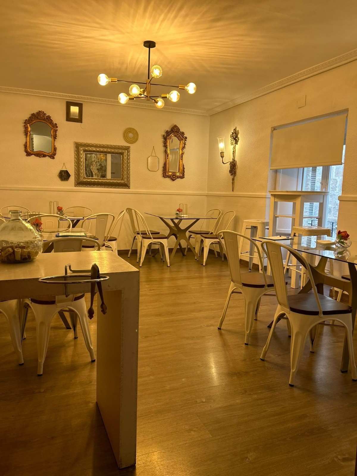 Comedor con iluminación cálida, sillas blancas, mesas de madera, espejos y grandes ventanales.