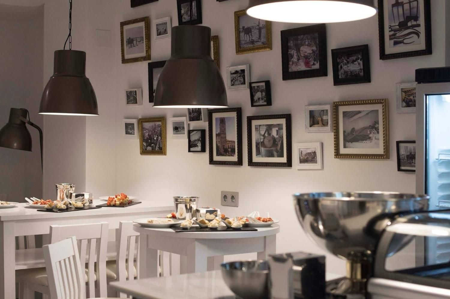 Interior de cafetería moderna con mesas blancas, lámparas colgantes y una pared de fotos enmarcadas.