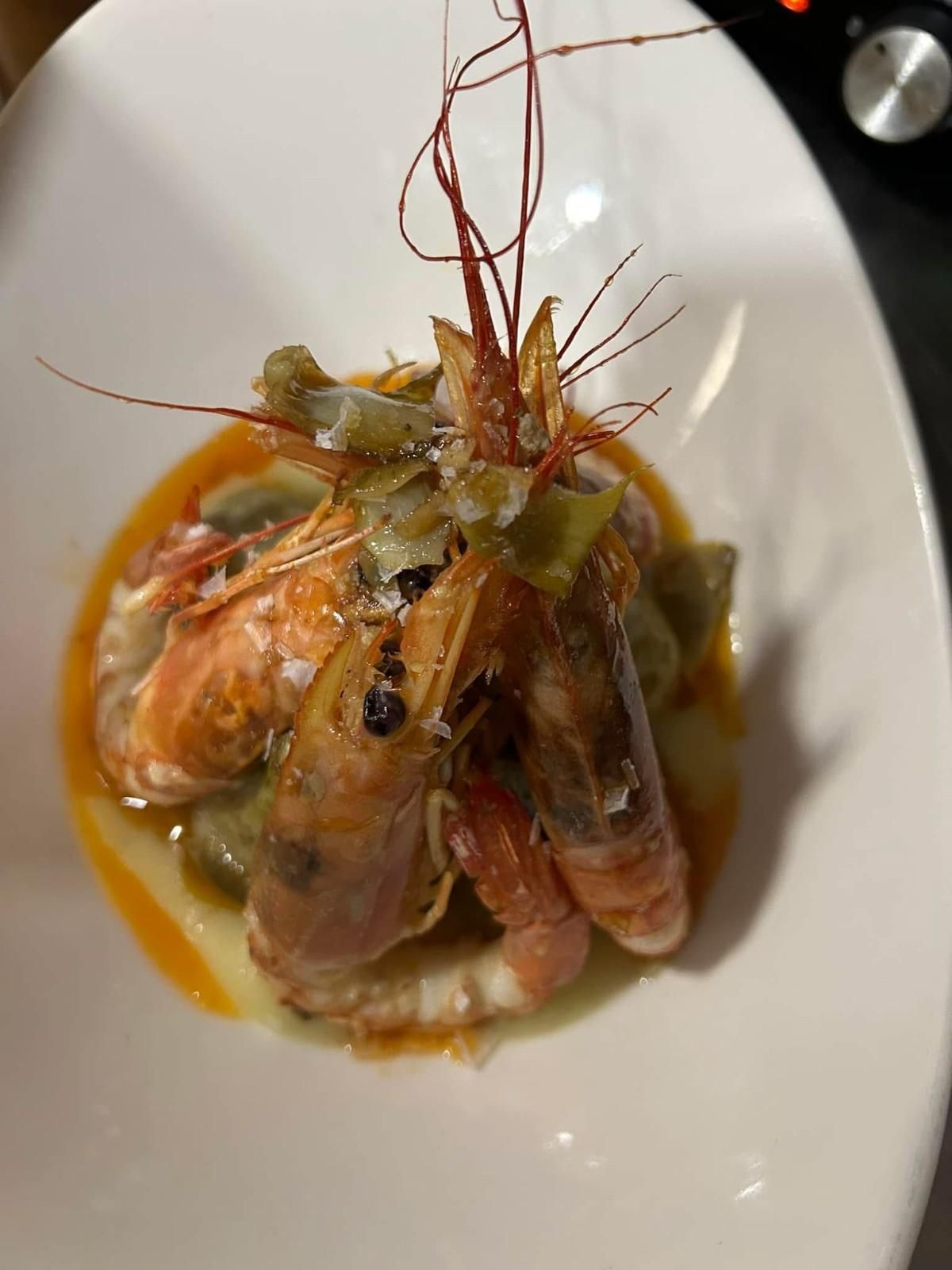 Plato gourmet de mariscos con langostinos en salsa sobre un plato blanco, adornado con hierbas y finas hebras crujientes.