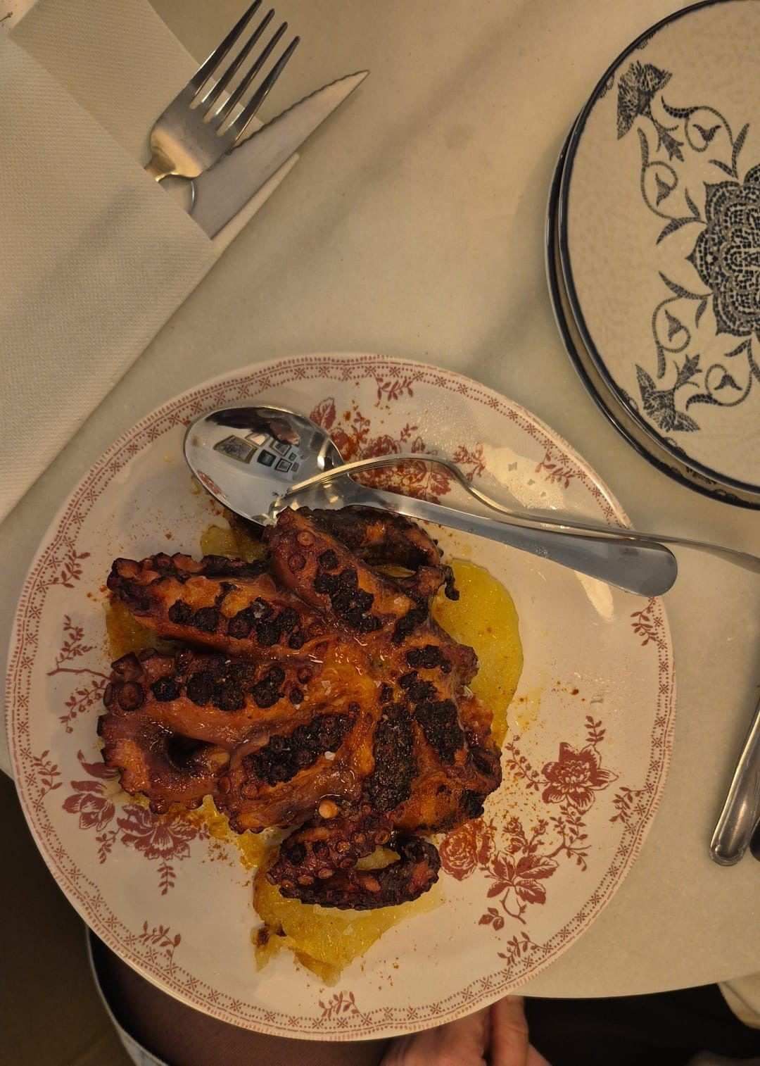 Comida a la parrilla emplatada con salsa en un plato decorado, con tenedores y cuchara en una mesa de restaurante.