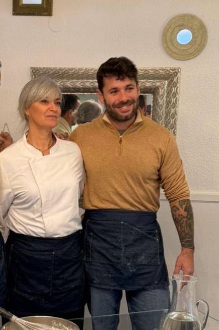Dos personas de pie juntas en un comedor, sonriendo junto a una mesa con una jarra de cristal.