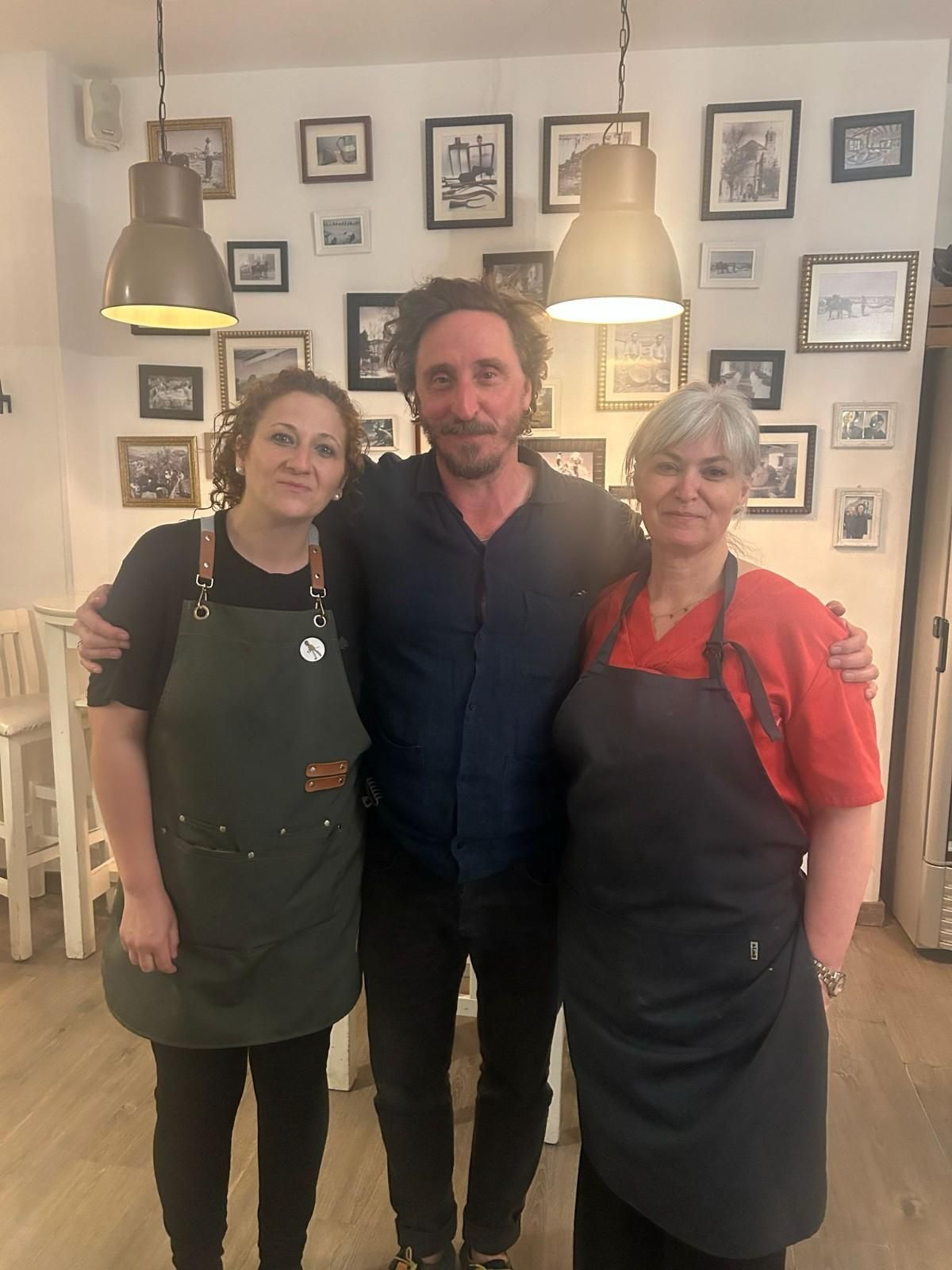 Tres personas de pie juntas en un acogedor café, sonriendo, con delantales puestos y fotos enmarcadas en la pared.
