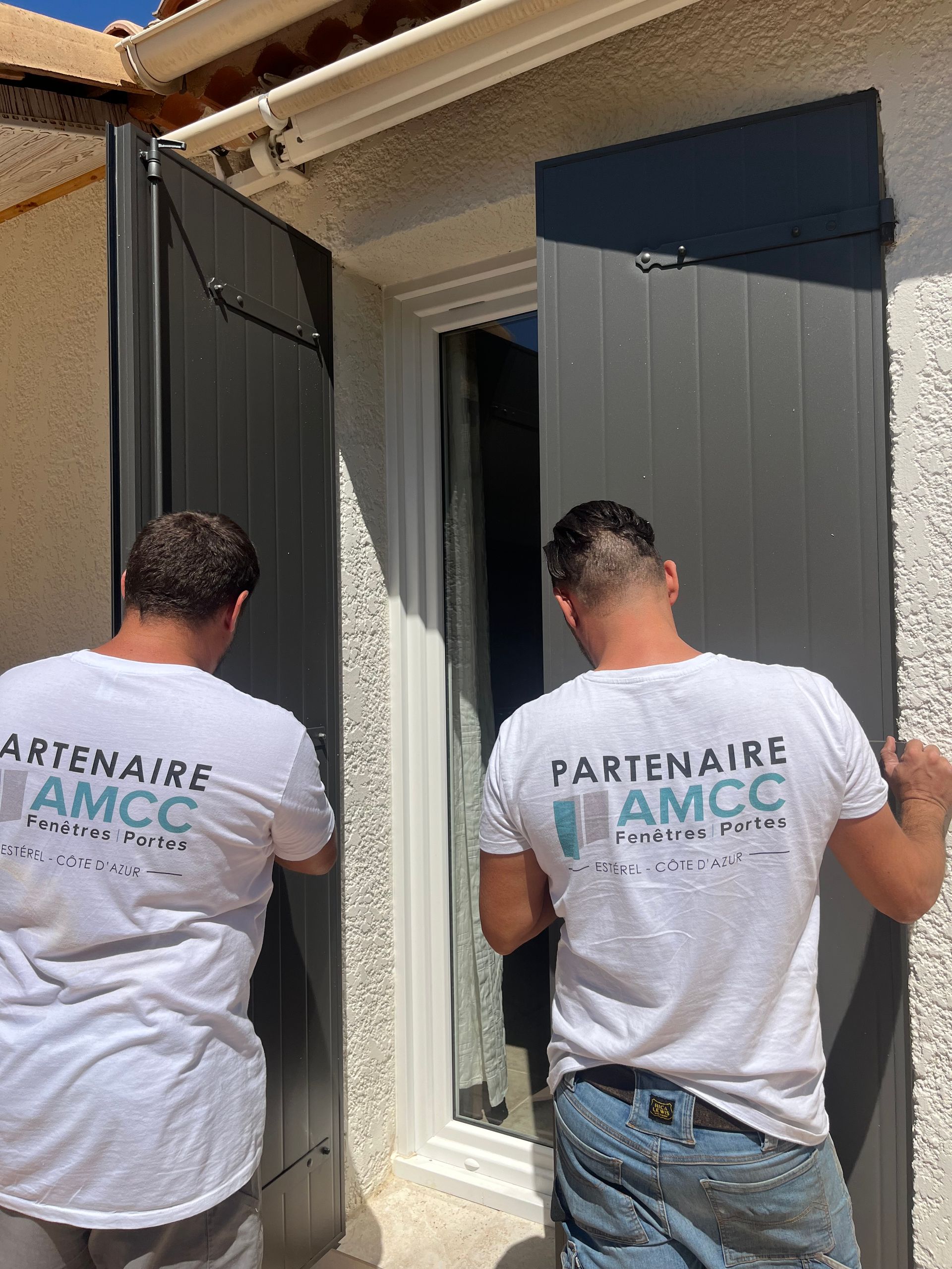 Deux techniciens de l'entreprise AMCC Fenêtres installent des volets gris sur la fenêtre d'une maison.