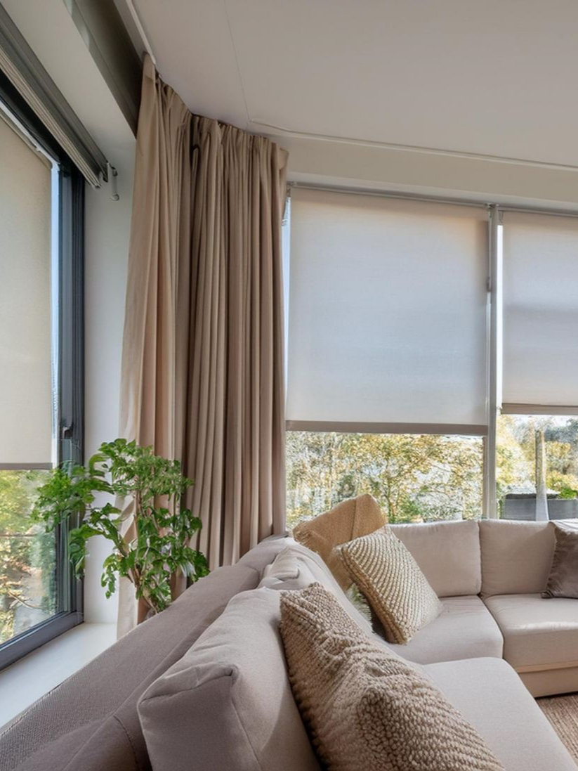 Un canapé d'angle beige est placé près de grandes fenêtres aux stores et rideaux aux tons neutres. Une plante est disposée près d'une fenêtre.