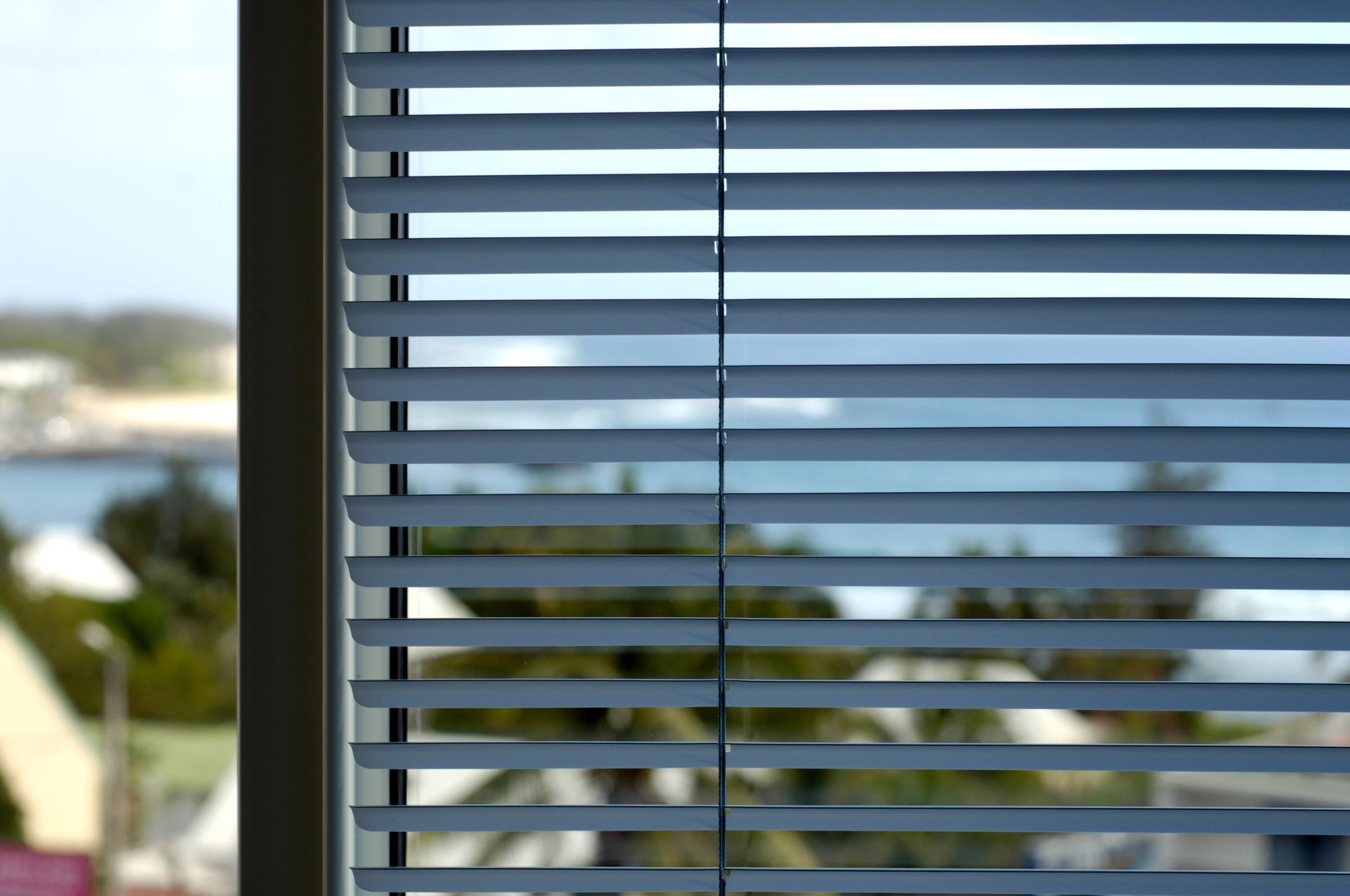 Des stores vénitiens masquent partiellement une scène extérieure floue avec des arbres et de l'eau.