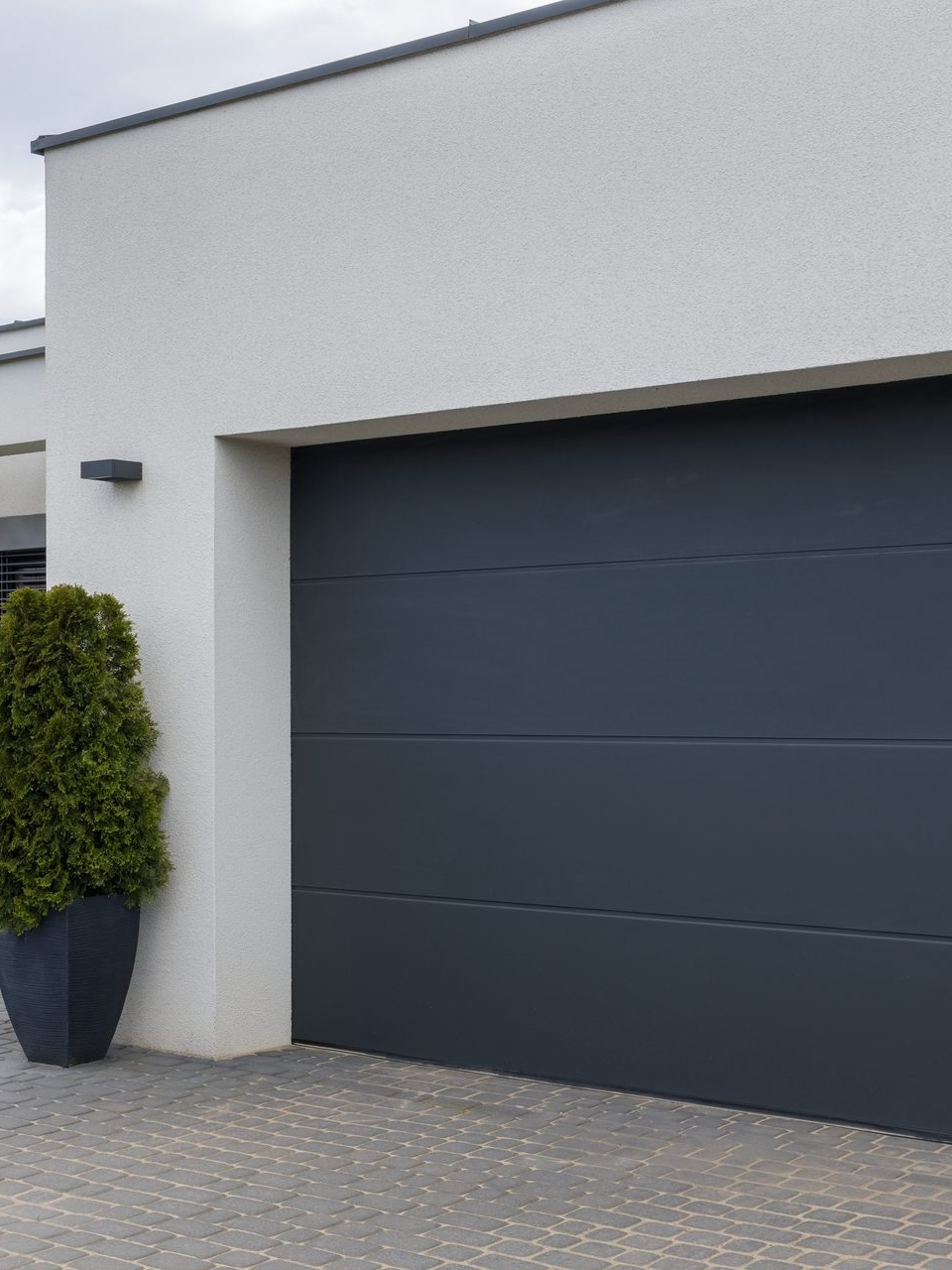 Porte de garage grise sur un bâtiment blanc, avec une plante dans un pot à gauche.