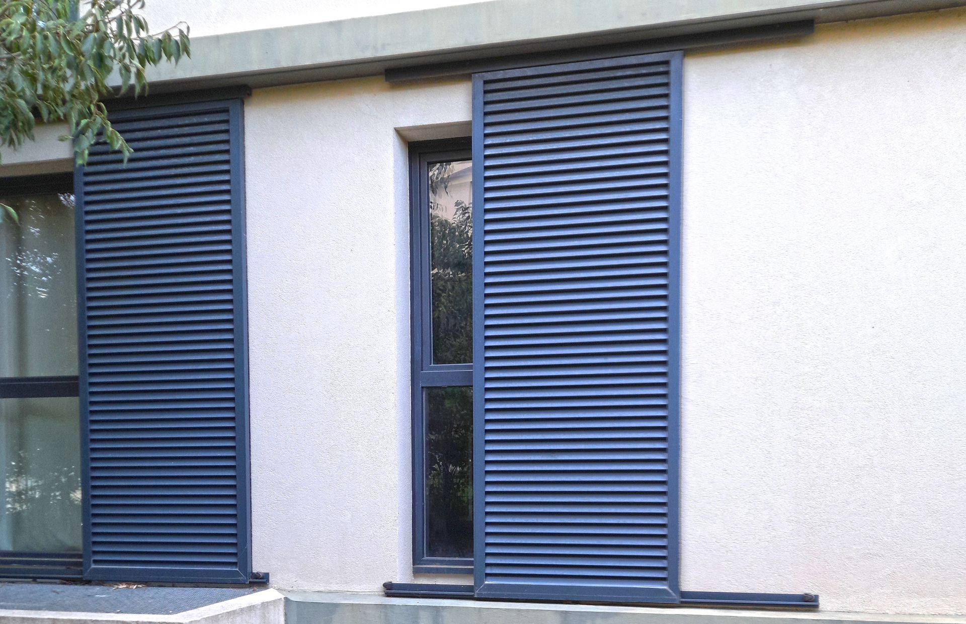 Des volets persiennes bleus coulissants s'ouvrent pour révéler la fenêtre d'une maison.