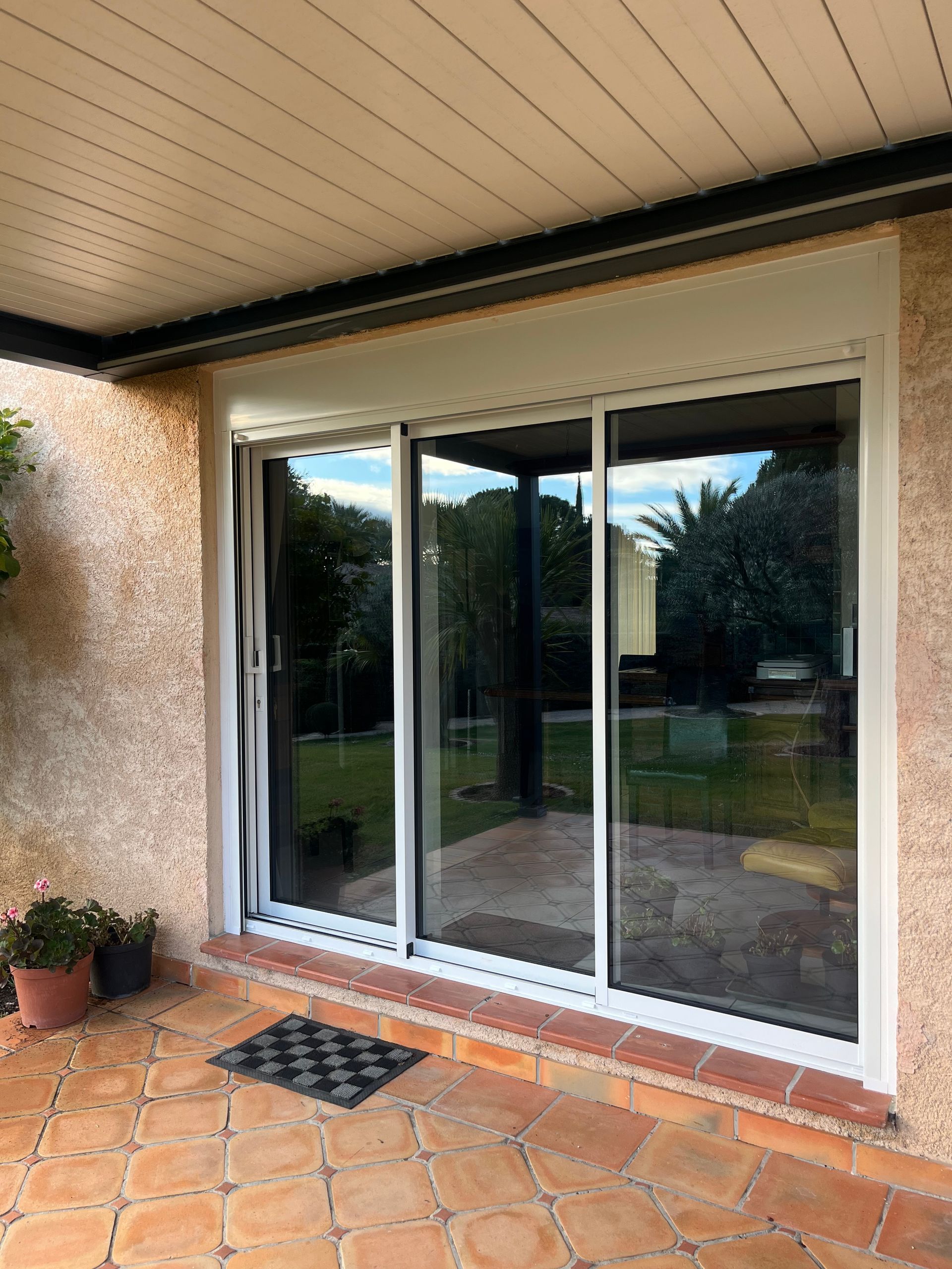 Baie vitrée à cadre blanc et volet enroulé donnant sur une terrasse carrelée, couverte d'une pergola bioclimatique.