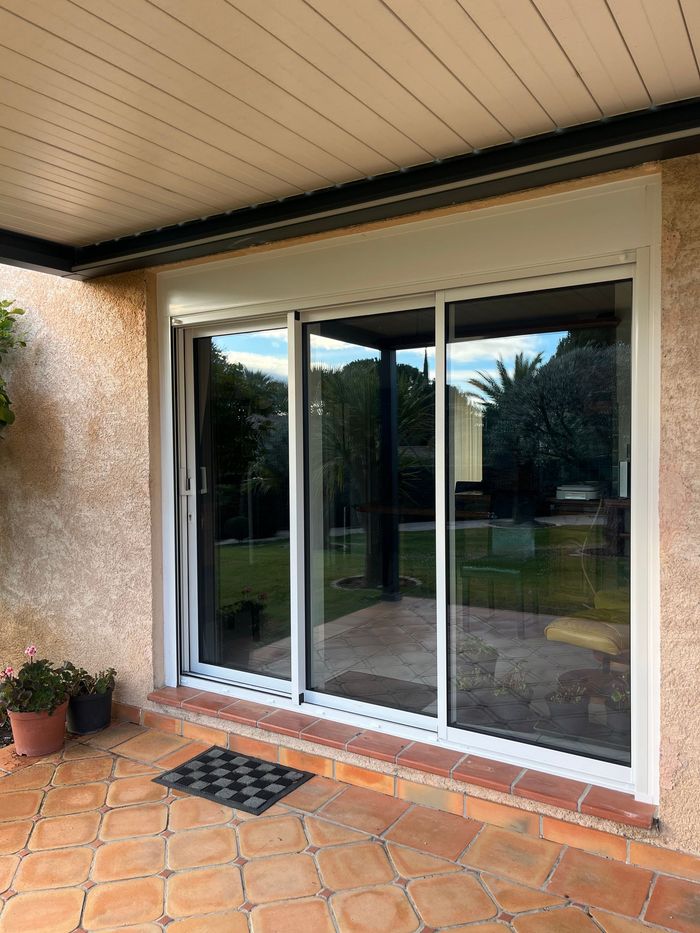 Baie vitrée à cadre blanc et volet enroulé donnant sur une terrasse carrelée, couverte d'une pergola bioclimatique.
