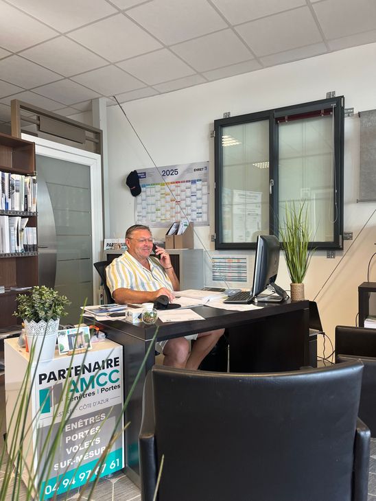 Sébastien de l'entreprise AMCC Fenêtres est au bureau, au téléphone, souriant. Son bureau est équipé d'un ordinateur, de documents et de plantes.